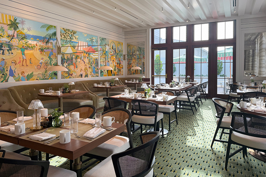 Bright, elegant restaurant with large windows, light green patterned carpet, wooden tables set for dining, cream chairs, and colorful beach-themed mural on the wall.