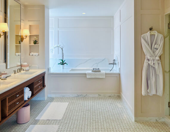 Luxurious bathroom with a double sink, bathtub, white robes hanging on the wall, white mats on a tiled floor, and an orchid plant by the tub. The space is bright and neatly organized.
