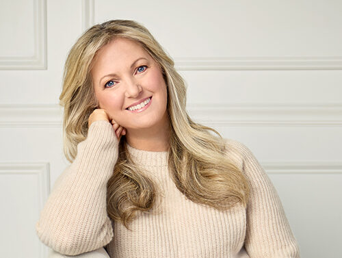 A woman with long blonde hair, wearing a beige sweater and white pants, sits on a cream chair, smiling and resting her head on her hand against a light-colored background.