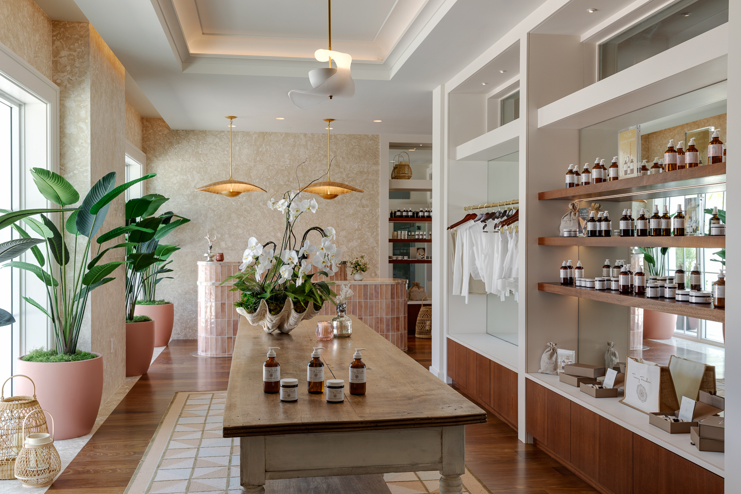 A modern, airy spa interior with wooden floors, potted plants, a central table displaying jars, shelves with bottles, and folded towels. Natural light streams in through large windows.