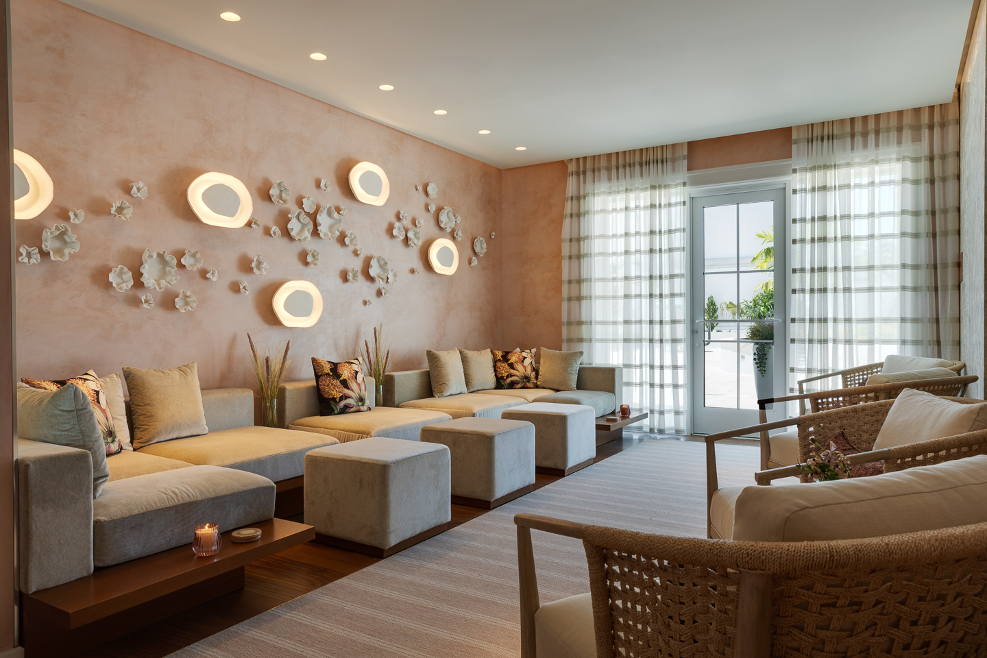 Modern lounge with beige sofas, light wood accents, decorative wall art with abstract lights, potted plants, woven chairs, and sheer curtains over a glass door leading to a sunny outdoor area.