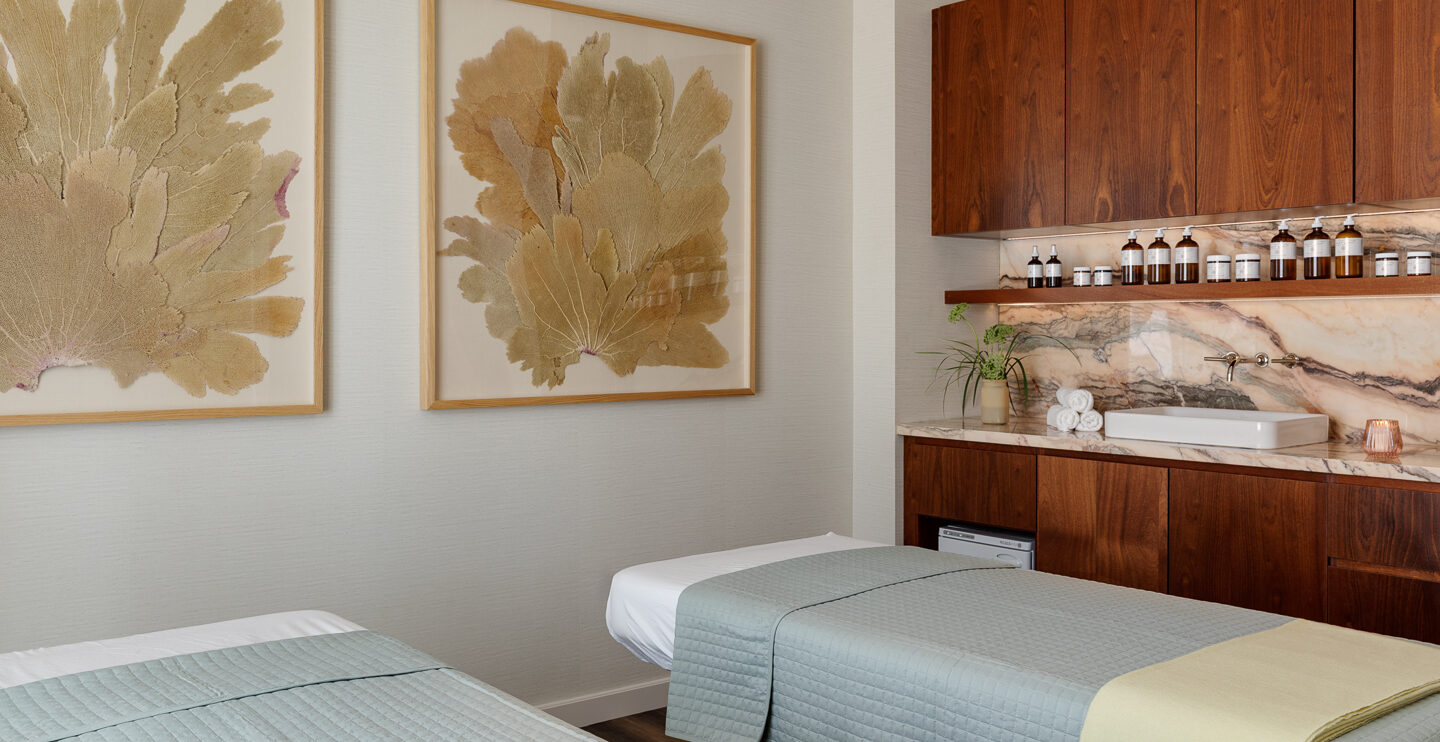 A modern spa room with two massage tables, green-gray quilts, a marble counter with a sink, wooden cabinets, and large framed botanical art on cream-colored walls. Bottles and towels are neatly arranged on the counter.