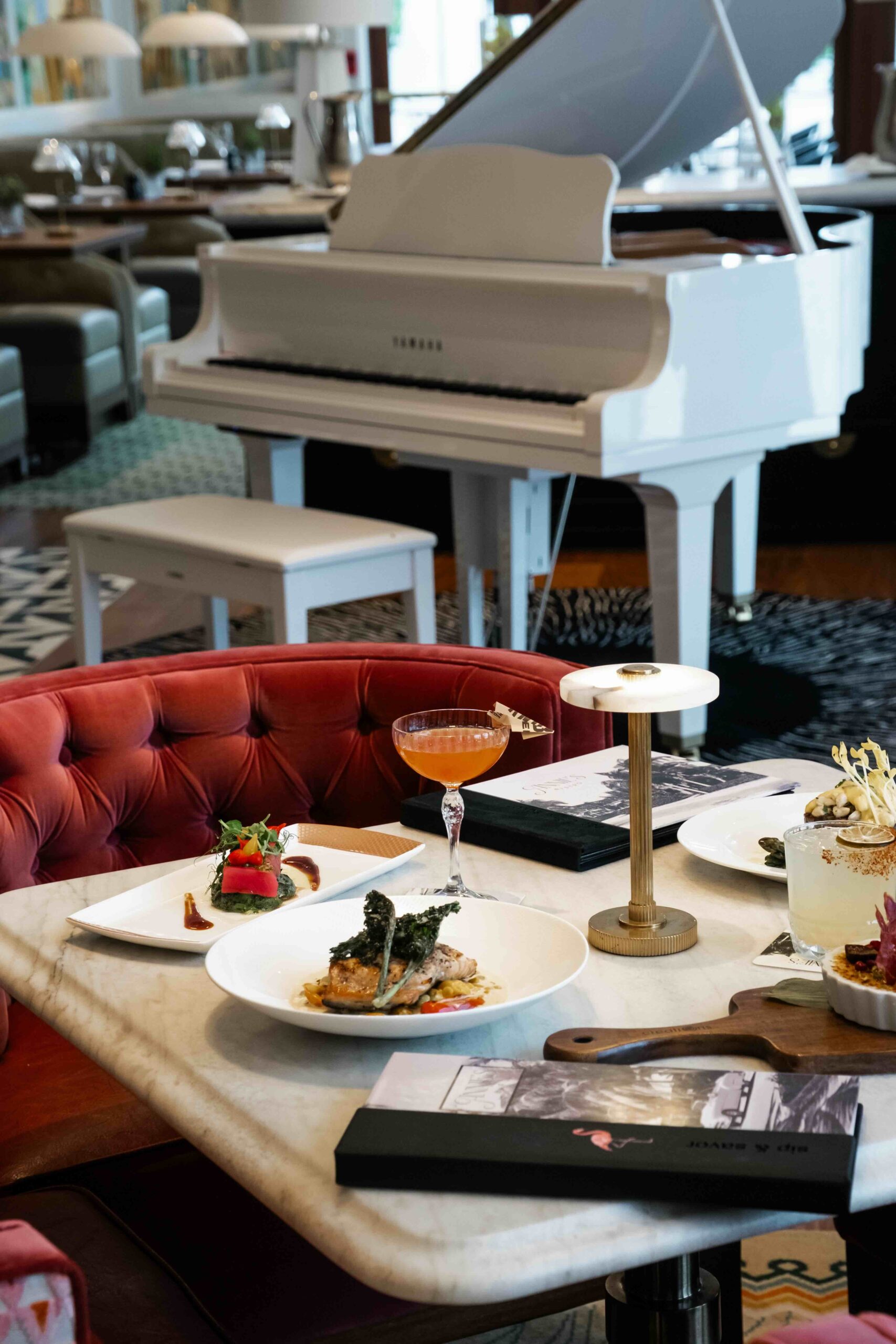 A marble table with plated gourmet food, drinks, and menus sits beside a red velvet chair in an elegant restaurant. A white grand piano is in the background.