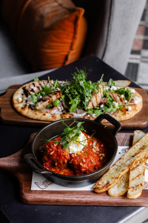 A skillet of tomato-based meatballs topped with cheese and herbs is served with breadsticks, alongside flatbreads with chicken, arugula, and balsamic glaze on a wooden board in a cozy restaurant setting.