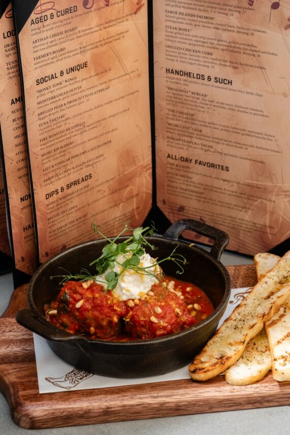 A black skillet with meatballs in tomato sauce, topped with cheese and microgreens, sits on a wooden board with toasted bread slices, with restaurant menus in the background.