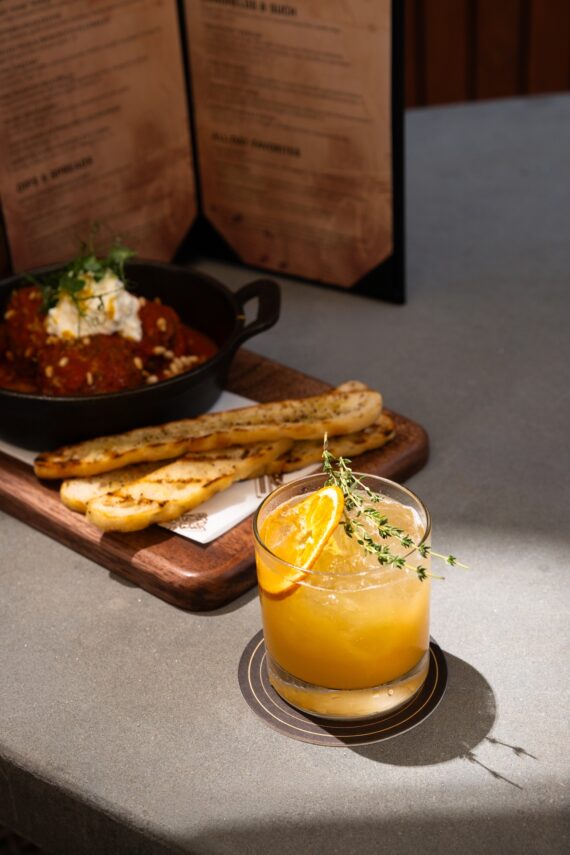 A cocktail garnished with an orange slice and herbs sits on a coaster in sunlight. Behind it, breadsticks and a skillet with dip rest on a wooden board, with a restaurant menu in the background.
