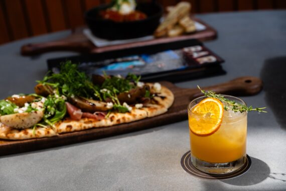 A flatbread topped with greens and vegetables sits on a wooden board, with a cocktail garnished with an orange slice and herbs in the foreground. The background is softly blurred.