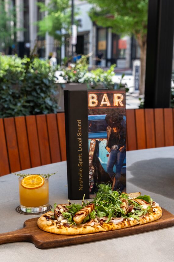 A flatbread pizza with greens and grilled vegetables on a wooden board, a cocktail garnished with an orange slice, and a BAR menu with a city street scene in the background.