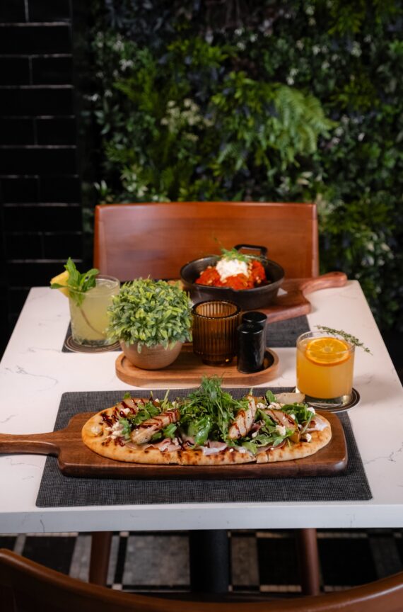 A table with a flatbread pizza, two colorful drinks, a potted plant, a candle, and a dish of meatballs with cheese, set against a leafy green background.