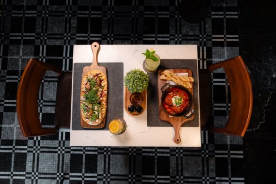 Overhead view of a restaurant table set for two, featuring flatbread, a skillet with tomato-based dish and bread, drinks, a small plant, and utensils on a checkered floor.
