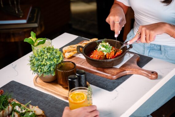 Person in a white shirt cutting into food in a black bowl on a wooden board, with drinks, bread slices, a potted plant, and salt and pepper shakers on a white table.