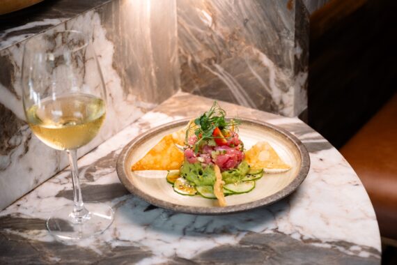 A glass of white wine sits next to a plate of tuna tartare with guacamole, cucumber slices, crispy chips, and microgreens on a marble countertop under a warm light.