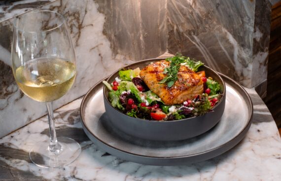 A bowl of salad topped with grilled salmon, cherry tomatoes, greens, feta, and pomegranate seeds sits beside a glass of white wine on a marble table.