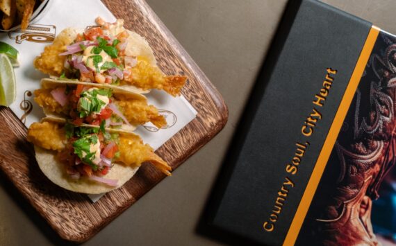 Two tempura shrimp tacos topped with pico de gallo and herbs sit on a wooden board next to a black book with Country Soul. City Heart written on the cover.