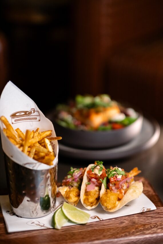 A serving of thin-cut fries in a metal cup, three fish tacos topped with salsa and herbs, and lime wedges on a wooden tray, with a blurred salad in the background.