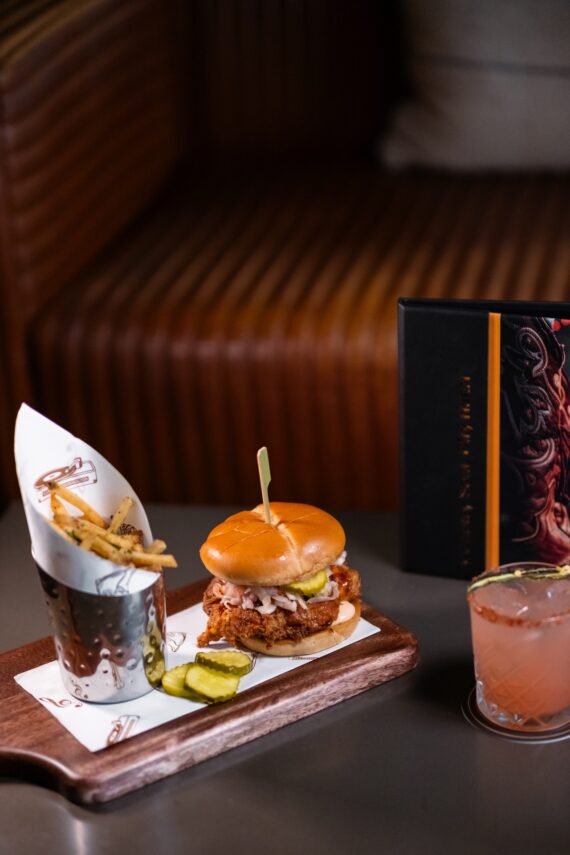 A fried chicken sandwich with pickles, a side of fries in a metal cup, and a pink cocktail sit on a wooden board, with a black menu in the background on a table near a brown booth.