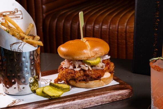 A fried chicken sandwich with pickles and slaw sits on a tray next to a metal cup of seasoned fries and a drink with a spiced rim.
