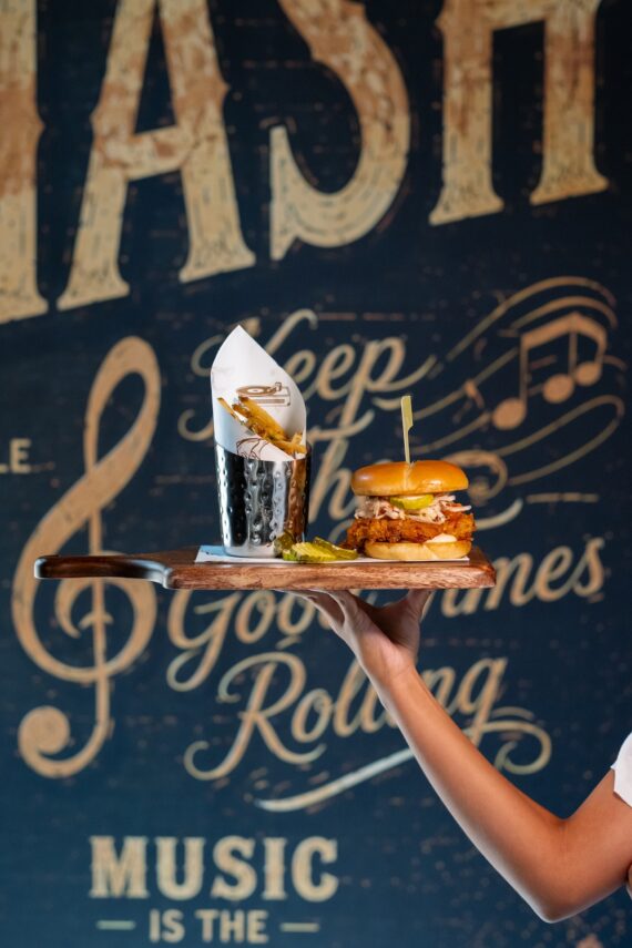 A hand holds a wooden tray with a burger, fries in a metal cup, and pickles. The background features a decorative wall with cursive text and musical symbols.
