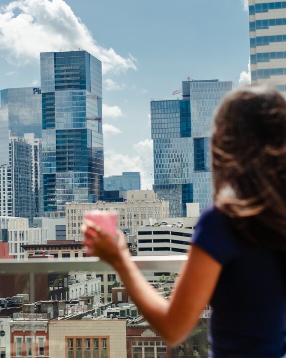 A woman with long hair, holding a pink drink, looks out at a cityscape with modern glass skyscrapers and older buildings under a blue sky with clouds.