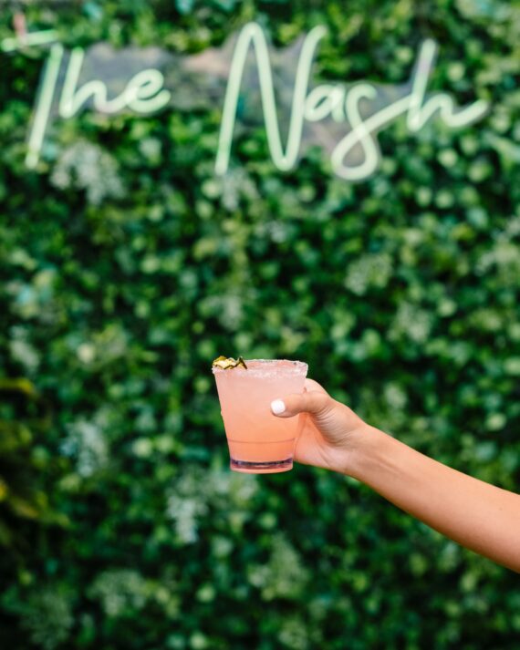 A hand with white nail polish holds a pink cocktail in front of a green leafy wall with The Nash written in neon script. The drink has a salted rim and a lime garnish.