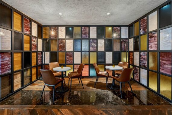 A modern lounge with four round tables, brown leather chairs, and colorful glass panels on the walls in shades of gold, amber, black, and white. The floor combines dark marble and wood parquet.