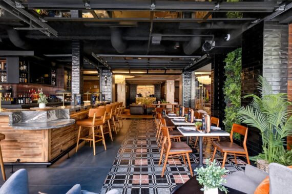 Modern restaurant interior with wooden tables and chairs, bar area on the left, patterned floor tiles, green plants, and warm lighting, creating a welcoming and stylish dining atmosphere.