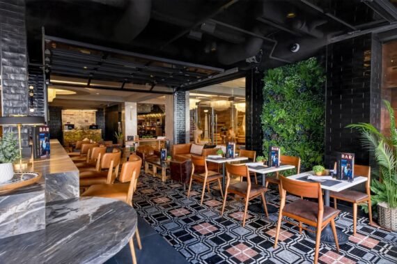 Modern restaurant interior with wooden tables and chairs, a green plant wall, and patterned tile flooring. Menus are on each table, and large windows let in natural light, creating a welcoming atmosphere.