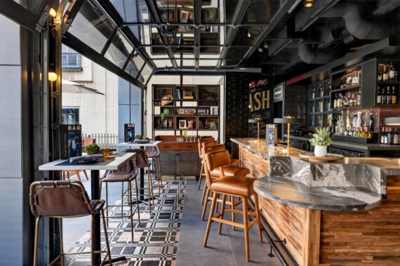 Modern bar with brown leather stools, a marble-topped counter, shelves with bottles and decor, and tall tables by large open windows. Sunlight brightens the stylish, inviting interior with industrial and wood accents.