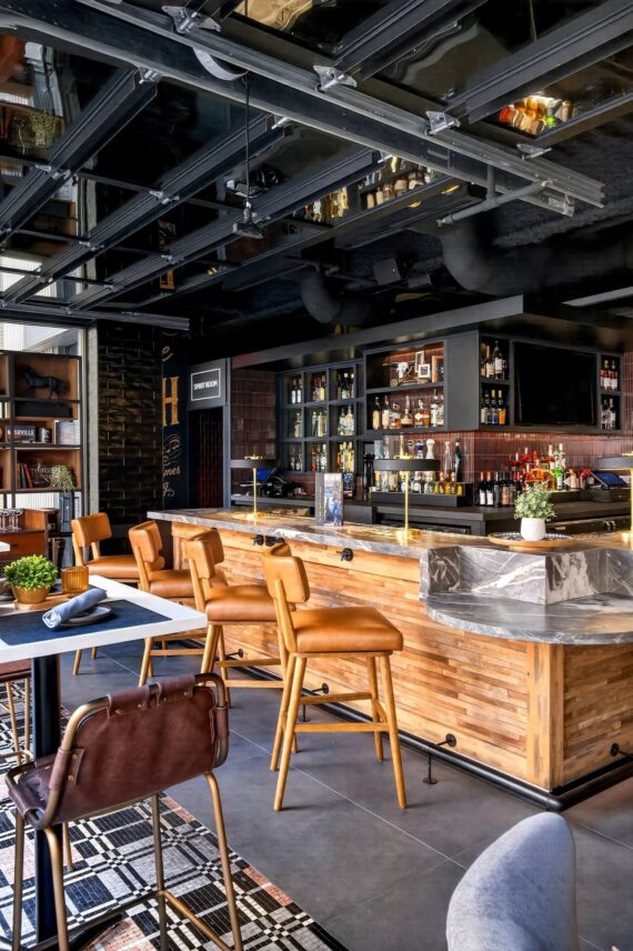 Modern bar with leather barstools, wooden counter, shelves stocked with bottles, and a mix of dining tables and chairs. Industrial decor with a cozy, inviting atmosphere and contemporary accents throughout the space.