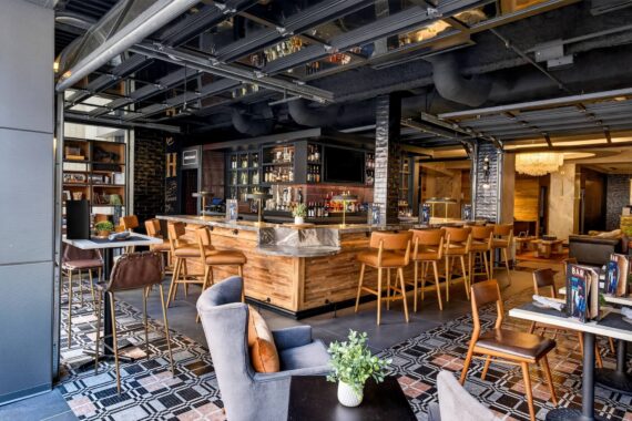 Modern restaurant interior with wooden bar, high chairs, lounge seating, patterned tile floor, and decorative plants. Shelves with bottles and glasses are behind the bar, and large windows let in natural light.