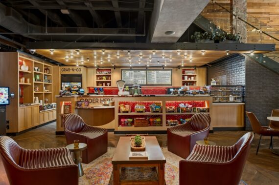 A cozy café interior with four brown leather chairs, a low table, and shelves stocked with snacks. The counter in back has a menu board, lights above, and a GRAB-N-GO sign. Warm lighting creates an inviting atmosphere.