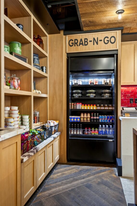 A hotel pantry with shelves of snacks, cereals, and drinks, and a refrigerator labeled GRAB-N-GO stocked with bottled beverages, sandwiches, and other quick food items.