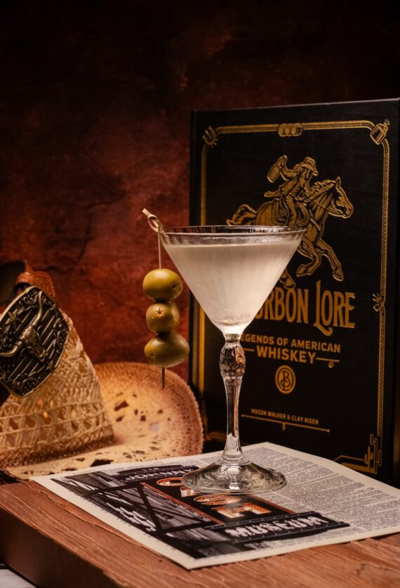 A martini with three olives on a skewer sits on a newspaper, next to a straw hat and a book titled Bourbon Lore: Legends of American Whiskey against a rustic brown background.