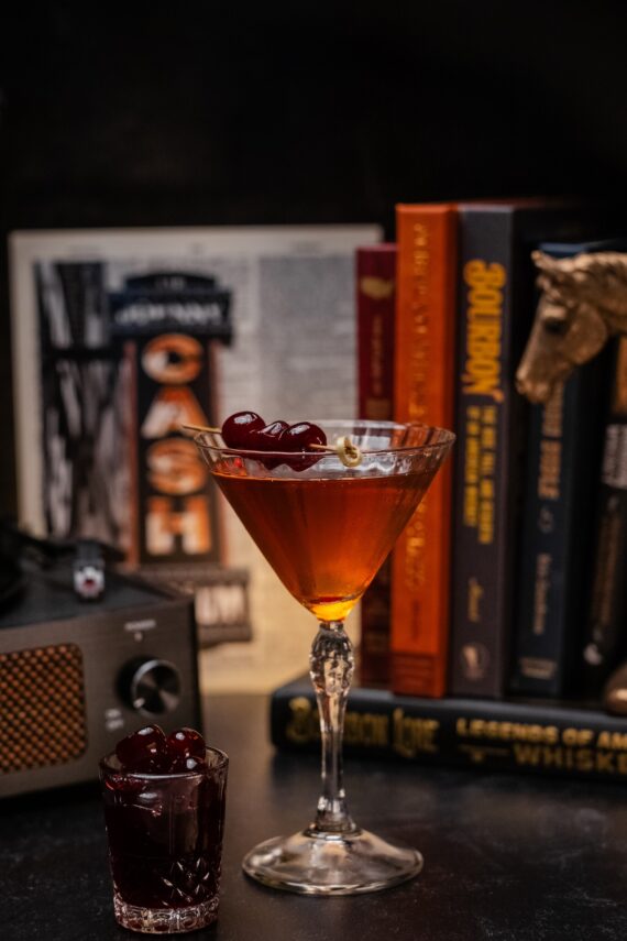 A cocktail in a martini glass garnished with cherries sits on a table next to a small glass of more cherries, with books and a vintage radio in the background.