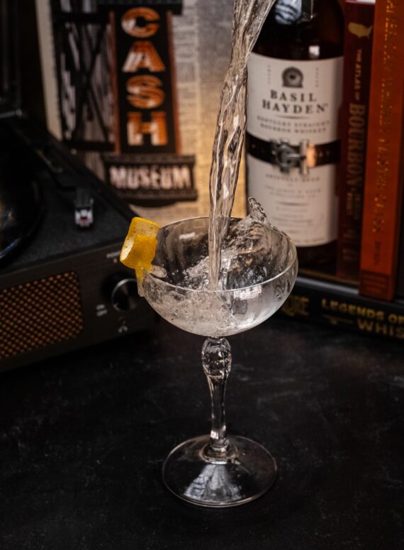 A clear liquid is being poured into a coupe glass with a lemon twist garnish, causing a splash. Bottles, books, and a record player are visible in the background.