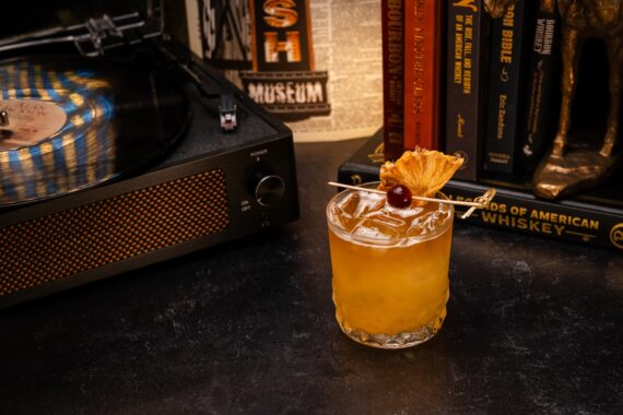 A cocktail garnished with dried pineapple and a cherry sits on a dark surface next to a record player and books, including titles about whiskey.