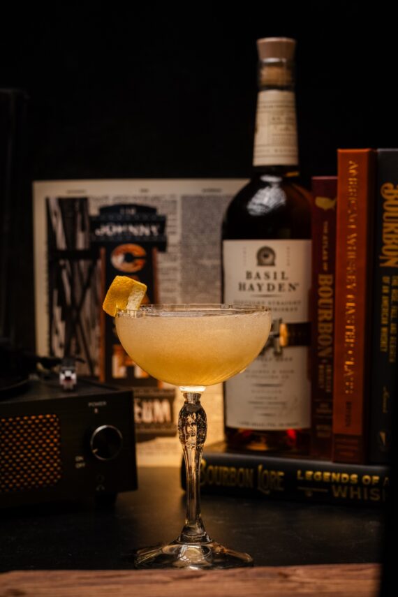 A coupe glass with a light yellow cocktail garnished with an orange peel sits in front of a Basil Hayden bourbon bottle, surrounded by books and a vintage radio on a dark background.