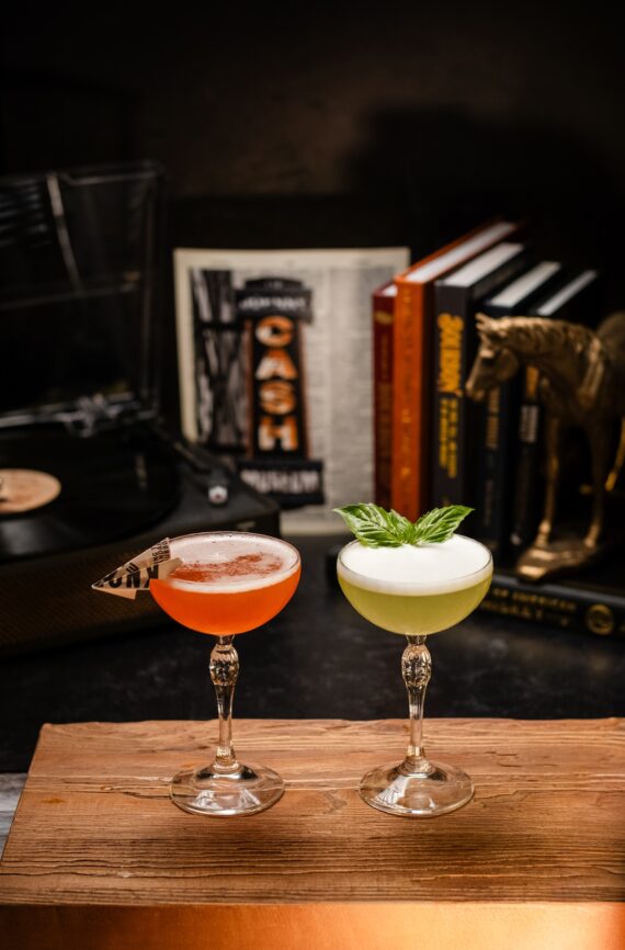 Two elegant cocktails on a wooden surface: one orange with a butterfly garnish, the other green with a basil leaf garnish. Books, a horse statue, and a record player are in the background.