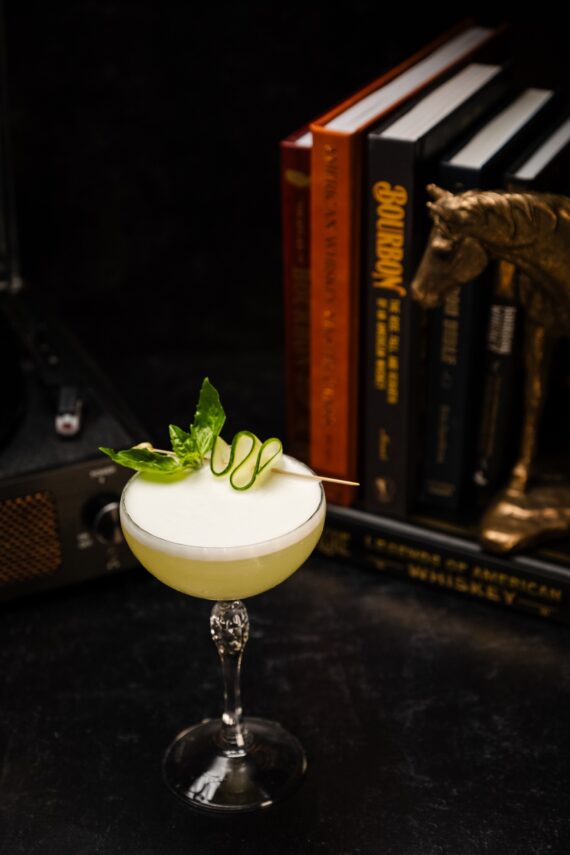 A cocktail garnished with cucumber and basil sits on a dark surface, in front of stacked books and a decorative horse head.