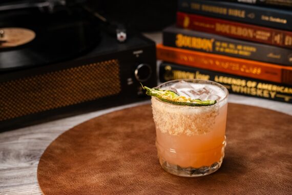 A cocktail with a salted rim and a green garnish sits on a brown mat. In the background, theres a stack of books about bourbon and whiskey next to a vintage record player.