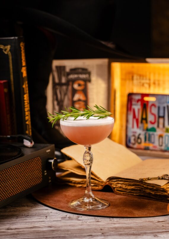 A pink cocktail with a frothy top, garnished with rosemary, in a stemmed glass on a table with an open book, vintage objects, and framed art in the background.