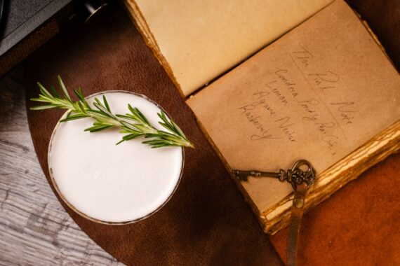 A frothy cocktail garnished with rosemary sits beside an old open book and a vintage key on a leather surface.