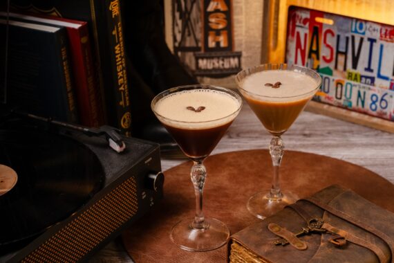 Two espresso martinis on a brown surface, each garnished with three coffee beans. Nearby are books, a record player, keys on an old book, and a colorful Nashville license plate in the background.