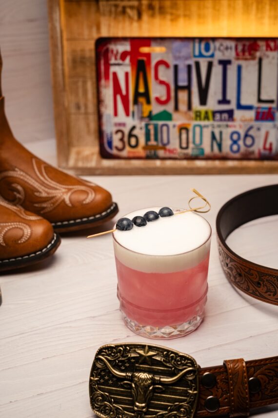 A pink cocktail with frothy top and blueberries is on a table, surrounded by cowboy boots, a tooled leather belt with a buckle, and a Nashville license plate in the background.