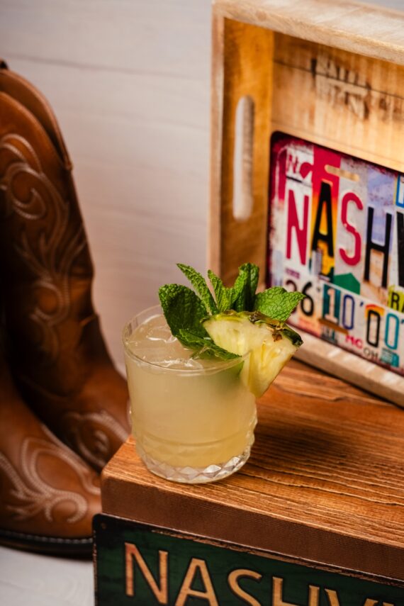 A cocktail garnished with pineapple and mint sits on a wooden box labeled Nashville, next to cowboy boots and a wooden crate with a decorative Nashville license plate.