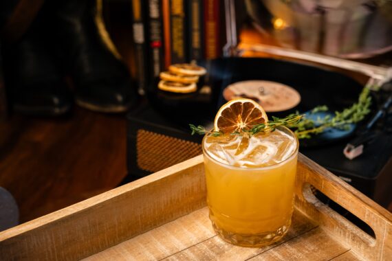 A yellow cocktail with ice, garnished with a dried orange slice and herbs, sits on a wooden tray. A record player, vinyl, books, and lemon slices are in the blurred background.