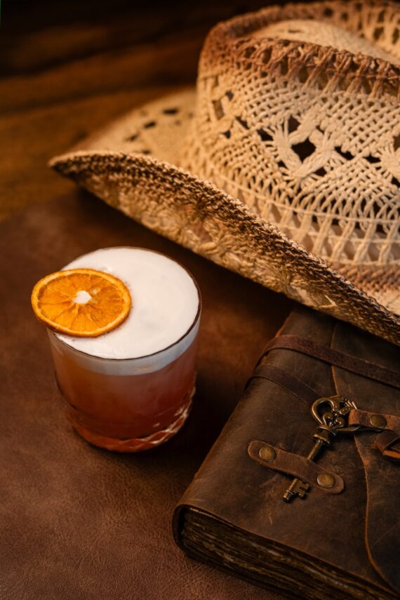 A cocktail with a frothy top and orange slice garnish sits on a leather surface beside a woven cowboy hat and an old leather-bound book with a key tied to it.