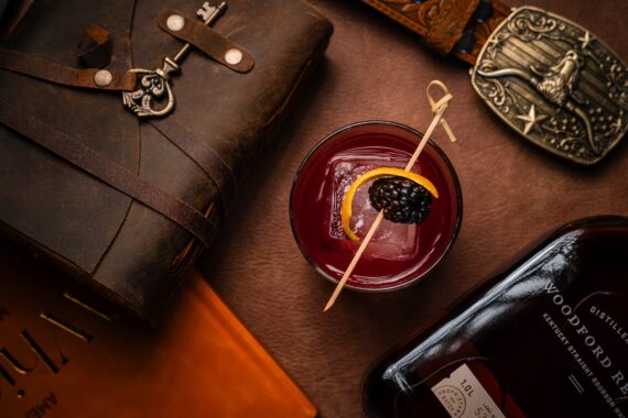 A cocktail garnished with an orange slice and blackberry sits on a leather surface, surrounded by a leather journal, a book, a belt with a western buckle, and a bottle of Woodford Reserve bourbon.