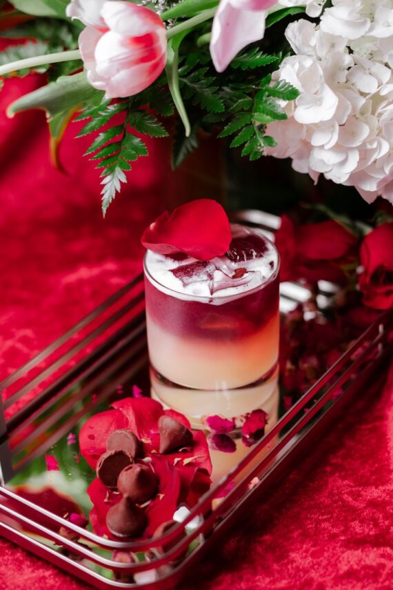 A layered cocktail garnished with red rose petals sits on a mirrored tray with chocolate truffles and scattered petals, surrounded by red and white flowers on a red velvet surface.