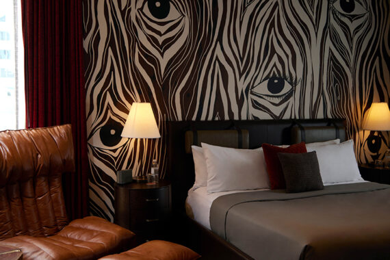 A stylish hotel room with a bed, brown leather chair, bedside tables with lamps, and a bold black-and-white wallpaper featuring zebra stripes and large eye designs. Red curtains hang beside a window.